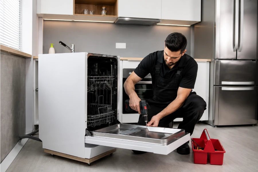 Appliance technician using a cordless drill on the lower panel of an open dishwasher pulled out from a kitchen cabinet run